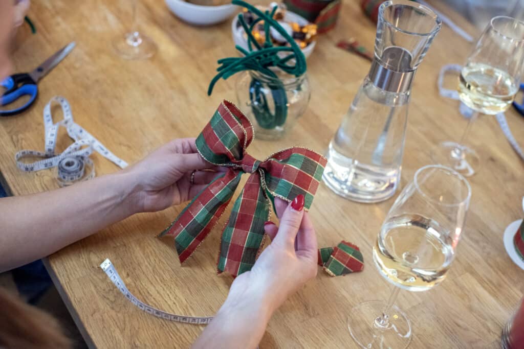 Person skapar julrosett vid ett bord, omgiven av hantverksmaterial och glas med dryck.